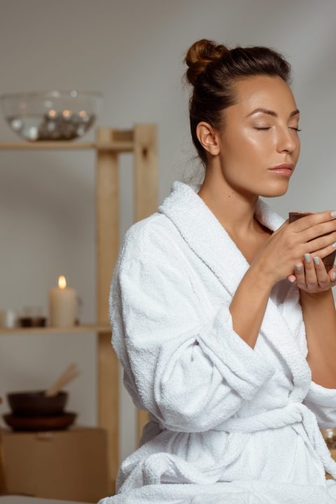 Young beautiful brunette girl holding tea cup relaxing in spa salon. Copy space.